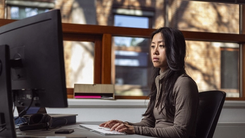 En kvinde sidder ved et skrivebord og arbejder ved en computer, iført en grå trøje og med mørkt, langt hår. Baggrunden viser store vinduer med naturligt lys, der strømmer ind i rummet.